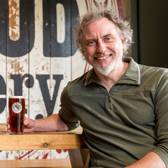 The Founder Greg, holding a beer at Stroud Brewery with a wooden bench and rustic wall in the background.