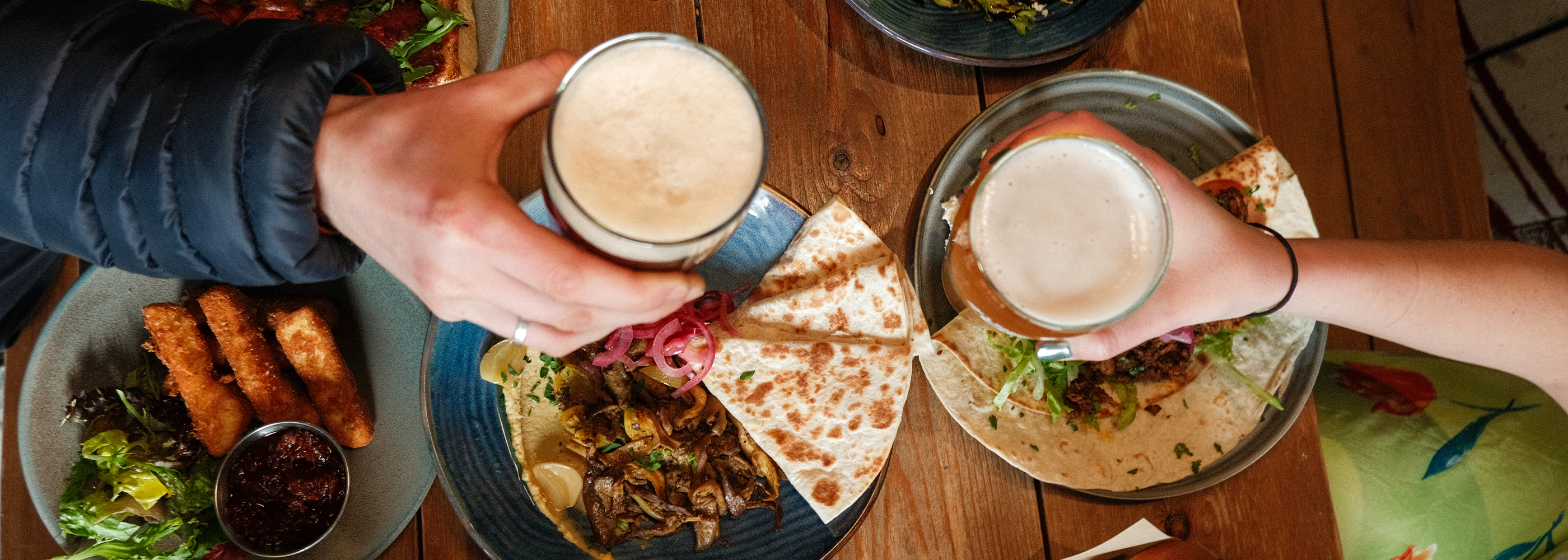 Two people holding drinks over plates of food on a wooden table