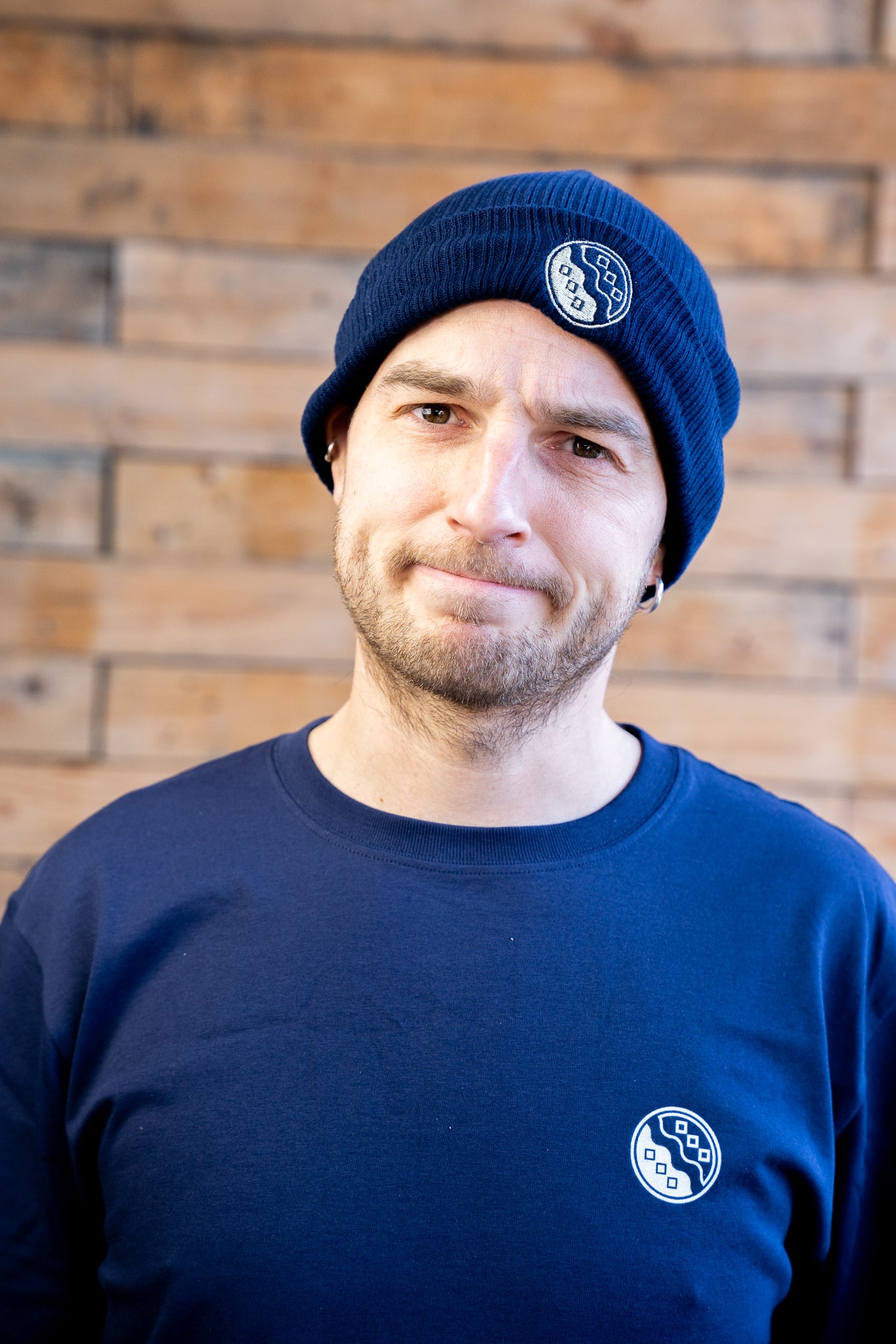 Man wearing a blue beanie and sweatshirt with a logo against a wooden wall.